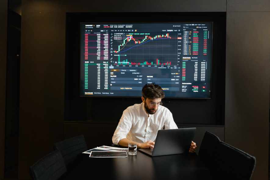 börskurser syns på stor skärm bakom en man som tittar på sin laptop